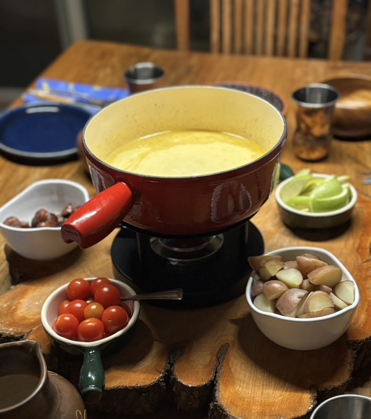 a Kuhn Rikon fondue pot filled with cheese fonde, and surrounded by small plates of food, including tomatoes, potatoes, mushrooms and green apples
